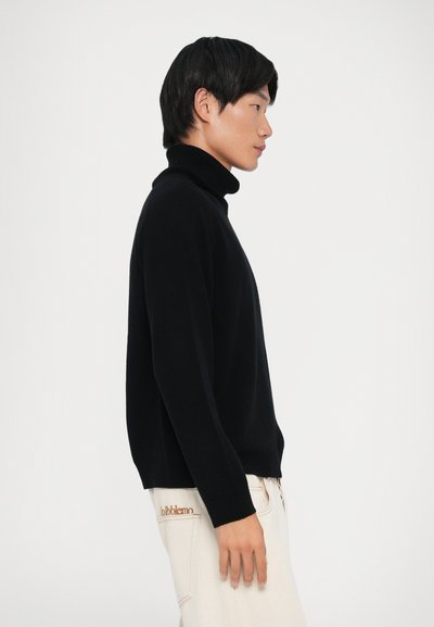 Maglione nero a collo alto con vestibilità morbida, maniche lunghe e dettagli a coste sul collo e sui polsini, abbinato a pantaloni di colore chiaro.