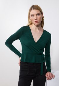 Zeleni crop top s dugim rukavima, V-izrezom i mašnom u struku. Glatka, rastezljiva tkanina u kombinaciji s crnim trapericama. Jednostavan dizajn.