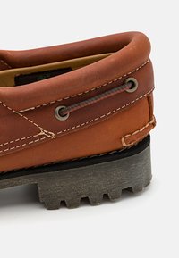 Brown leather shoe featuring a rounded toe, contrasting beige stitching, eyelets with braided laces, and a textured rubber sole.