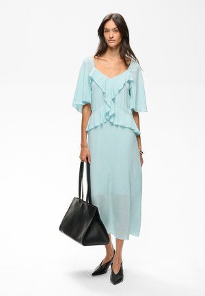 Femme portant une robe midi bleu clair à volants, tenant un grand sac à main en cuir noir et portant des chaussures pointues noires, debout sur fond blanc.