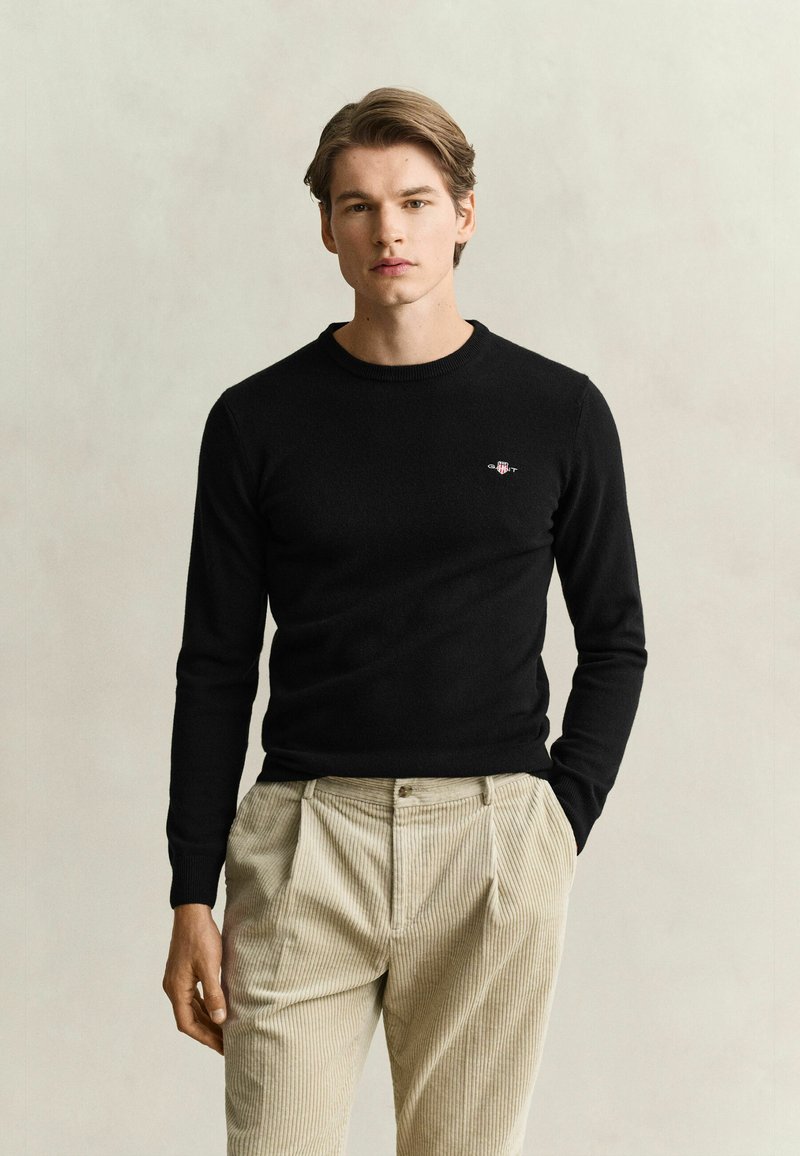 Jeune homme portant un pull noir à manches longues et un pantalon beige en velours côtelé, debout devant un fond clair uni avec une main dans la poche.