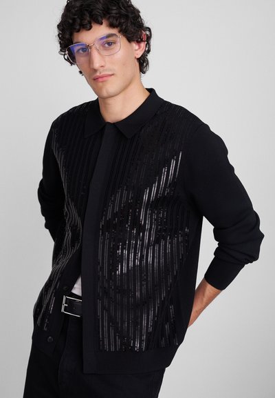 Cardigan noir avec un col structuré, présentant des rayures noires en sequins sur le devant, ainsi que des poignets et un ourlet côtelés.