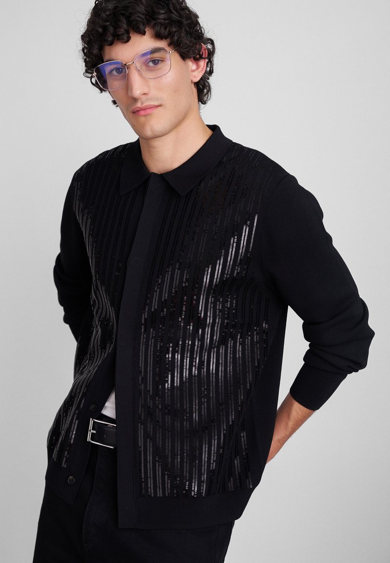 Cárdigan negro con un cuello estructurado, que presenta rayas de lentejuelas negras en la parte frontal y puños y dobladillo acanalados.