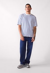 Camisa de rayas en tela blanca y azul, de manga corta, corte relajado. Combinada con vaqueros oscuros de pierna recta y zapatillas deportivas grises.
