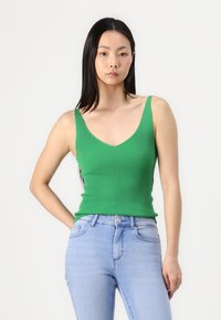 Green ribbed tank top with a deep V-neck and slim straps, paired with light blue skinny jeans, showcasing a fitted style.