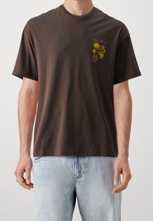 Uomo con una maglietta marrone scuro con piccolo motivo floreale giallo e testo giapponese rosso sul petto sinistro, abbinata a jeans azzurri.