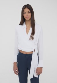 Blusa bianca avvolgente con maniche lunghe, scollatura a V e dettaglio con fiocco in vita, abbinata a pantaloni slim-fit blu navy. Texture del tessuto morbida.
