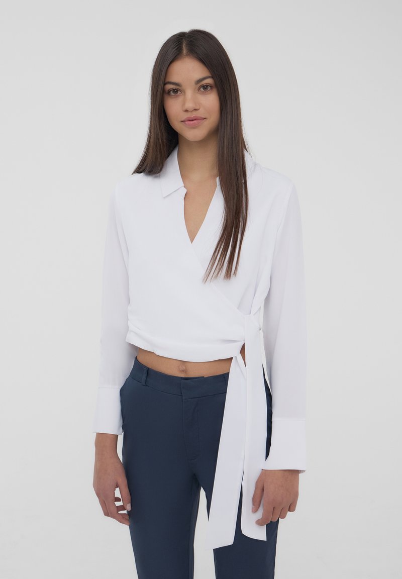 Blusa bianca avvolgente con maniche lunghe, scollatura a V e dettaglio con fiocco in vita, abbinata a pantaloni slim-fit blu navy. Texture del tessuto morbida.