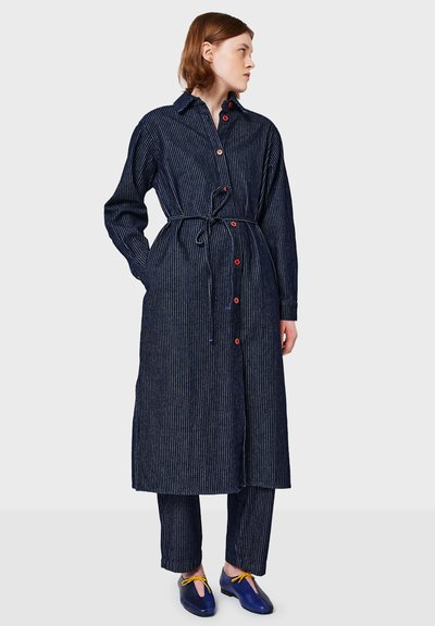 Kobieta w granatowej sukience koszulowej w prążki z czerwonymi guzikami, dopasowanych spodniach i niebieskich butach z żółtymi kokardkami, stojąca i patrząca w prawo.