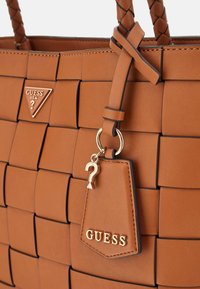 Borsa a tracolla intrecciata marrone con un logo triangolare, una tracolla intrecciata e un ciondolo staccabile a forma di punto interrogativo. Texture in pelle liscia.