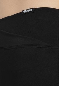 Zwarte leggings van een gladde, rekbare stof met een omgevouwen tailleband. Een genaaide label "SMILODOX" is zichtbaar op de tailleband.