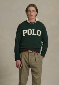 Maglione verde con "POLO" in lettere bianche, indossato sopra una camicia a strati; abbinato a pantaloni a quadri beige e una cintura. Dettagli testurizzati chiari.