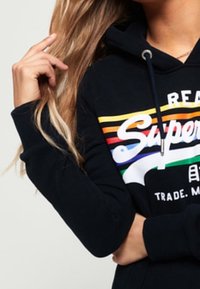 Donna che indossa un hoodie nero con un logo "Superdry" a strisce colorate, mentre tiene i suoi lunghi capelli biondi con una mano.