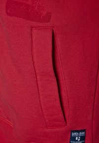 Sudadera roja con textura suave, que cuenta con un bolsillo frontal y una etiqueta de la marca con detalles en azul y blanco en la parte inferior derecha.