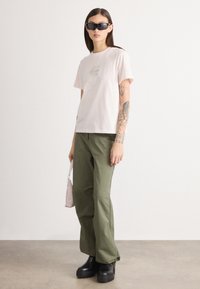 Ροζ κοντομάνικο μπλουζάκι με ένα μικρό γραφικό, συνδυασμένο με πράσινα φαρδιά παντελόνια και μαύρες πλατφόρμες μπότες; κρατώντας μια μικρή ροζ τσάντα.