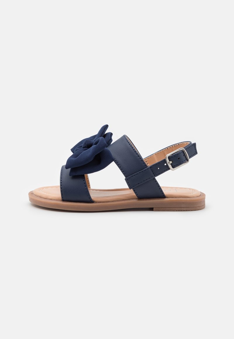 Sandal in blu navy con ampia tracolla, decorazione in tessuto a forma di fiore, cinturino alla caviglia regolabile e suola piatta in gomma. Texture liscia.