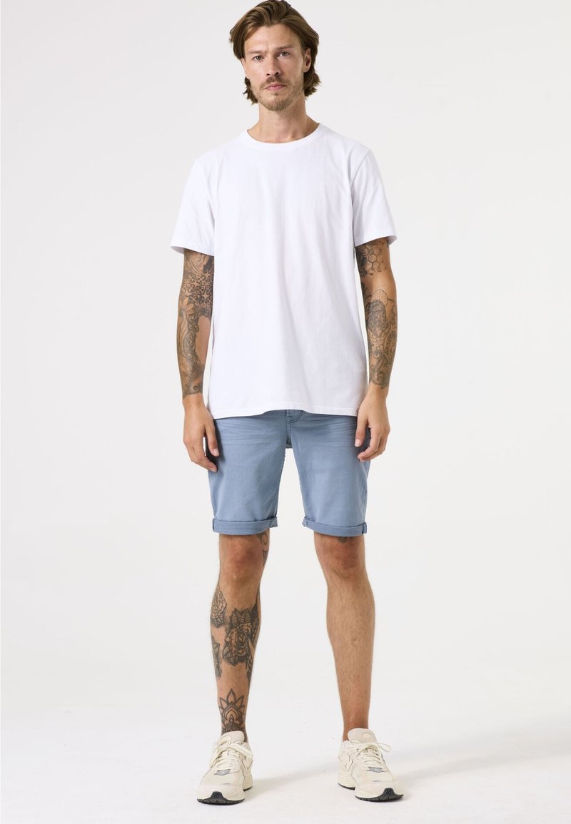 Camiseta blanca de algodón, pantalones cortos de mezclilla azul claro enrollados en los puños y zapatillas de crema. Tatuajes visibles en brazos y piernas. Fondo sencillo.