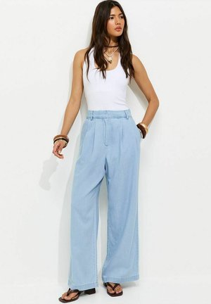 Femme portant un haut blanc sans manches, un pantalon large bleu clair, des sandales noires, des colliers superposés et des bracelets, debout contre un mur blanc.