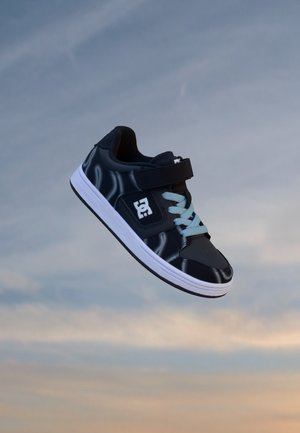 Baskets noires avec semelle blanche, lacets bleu clair et une bride à l'avant, flottant sur un fond de ciel nuageux.