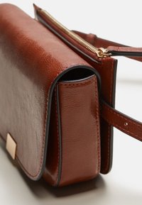 Brązowa skórzana torebka o gładkiej fakturze, prostokątnym kształcie, złotym suwadle i smukłym pasku. Posiada metalowy akcent z przodu.