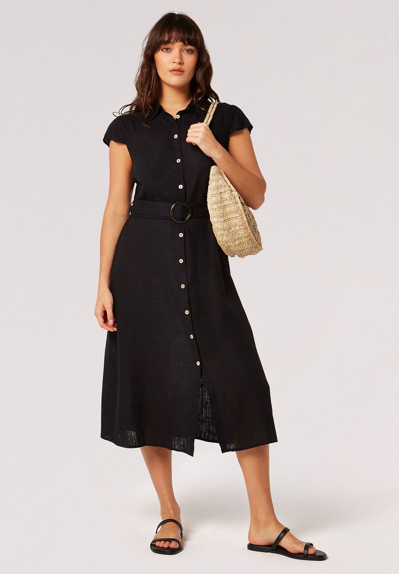 Schwarzes Knopfkleid mit kurzen Ärmeln, breitem Gürtel und gespaltem Saum. Hergestellt aus leichtem Stoff, kombiniert mit einer geflochtenen Tasche und Sandalen.