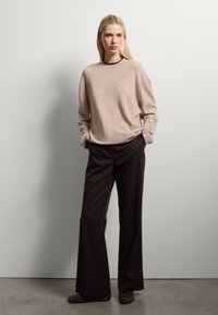 Pull léger beige à la coupe décontractée et au col rond, associé à un pantalon large marron foncé et des chaussures foncées, sur un fond neutre.