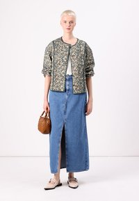 Groen gewatteerd jasje met bloemenpatroon en opbollende mouwen, gecombineerd met een denim maxirok met hoge split. Het model draagt een bruine handtas.