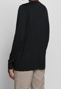 Pull noir à manches longues en maille avec texture côtelée et encolure arrondie, présentant un petit logo au dos. Porté avec un pantalon à carreaux.