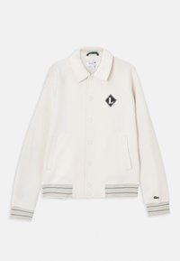 Veste blanche à boutons avec col, deux poches avant, poignets et ourlet rayés, et un emblème noir en forme de losange sur la poitrine gauche.