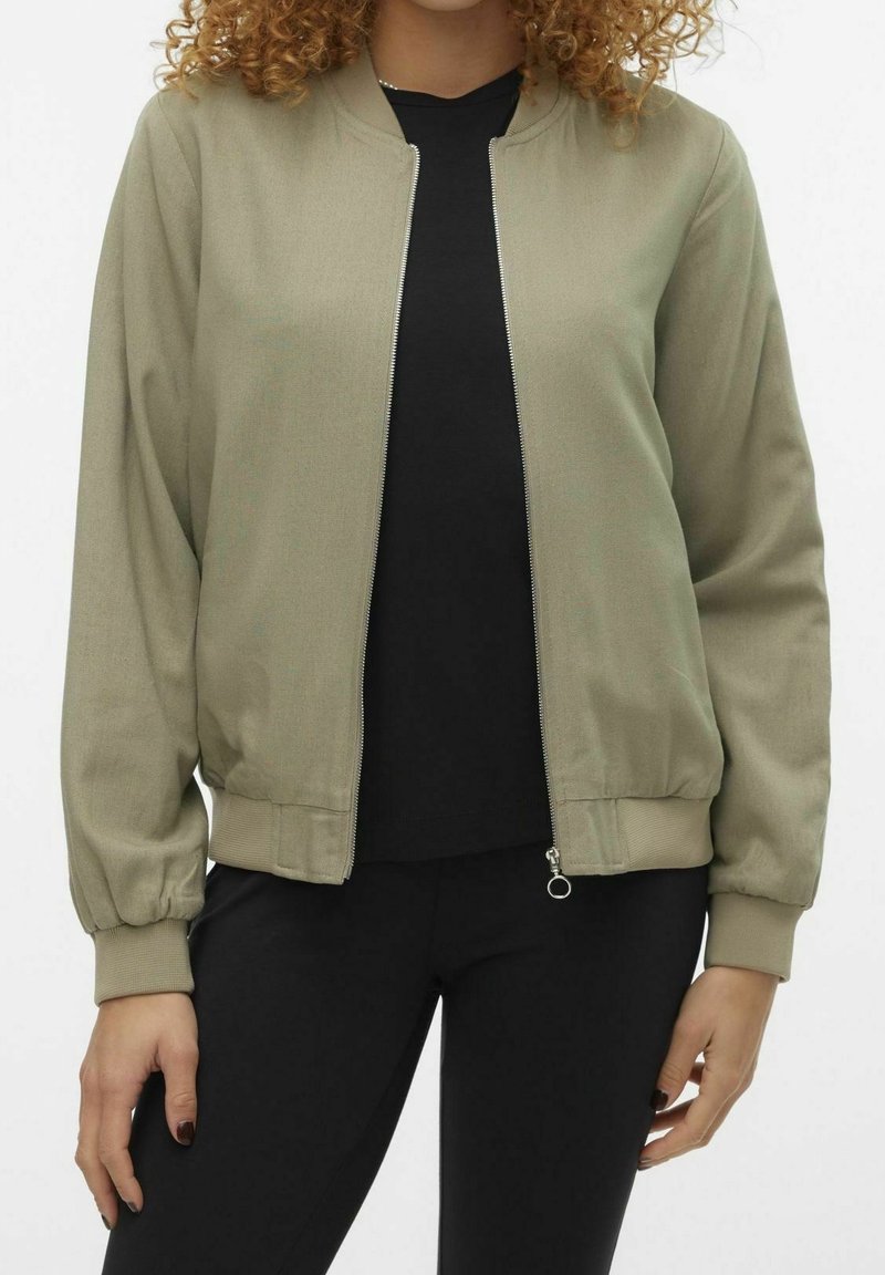 Jasnooliwkowa kurtka bomberka z zamkiem błyskawicznym, ściągaczami przy mankietach i u dołu. Wykonana z gładkiego materiału, zestawiona z czarną bluzką i spodniami.