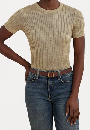 Personne portant un haut côtelé en maille dorée à manches courtes, rentré dans un jean bleu avec une ceinture marron, debout avec une main sur la hanche.