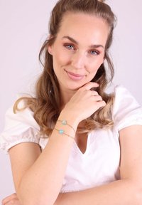 Gouden schakelarmband met twee turkooisstenen, één rond en één hartvormig, om een pols, tegen een effen achtergrond.