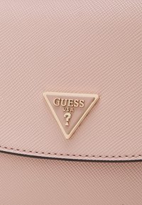 Superficie de cuero texturizado rosa con un logotipo triangular bordeado de oro que muestra "GUESS USA" y un signo de interrogación en el centro.