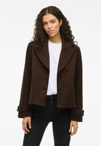 Joven mujer con cabello rizado que lleva una chaqueta oscura de botones sobre una blusa blanca y pantalones negros, de pie contra un fondo blanco.