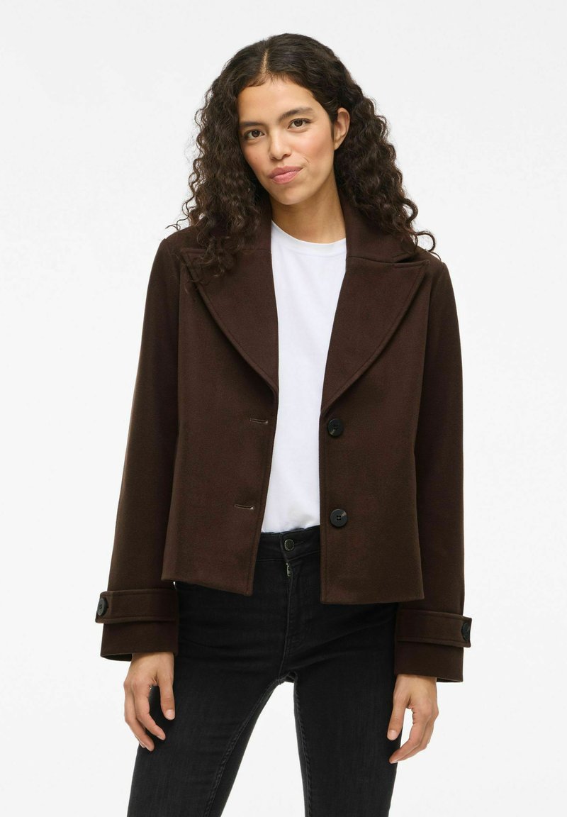 Joven mujer con cabello rizado que lleva una chaqueta oscura de botones sobre una blusa blanca y pantalones negros, de pie contra un fondo blanco.