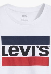 Camiseta blanca con un audaz logo negro de "Levi's" sobre un fondo rectangular azul y rojo. Diseño de cuello redondo con una suave textura de algodón.
