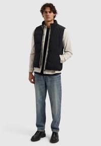 Zwarte puffer gilet over een lichtgrijze blouse, gecombineerd met donkerblauwe spijkerbroek en zwarte schoenen. Voorzien van een rits en ribbelstructuur.