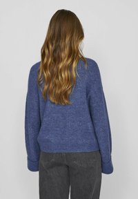 Pulover iz temno modre volnene mešanice, s sproščenim krojem, rebrastimi manšetami in mehko teksturo. V kombinaciji s temnosivimi jeans hlačami.