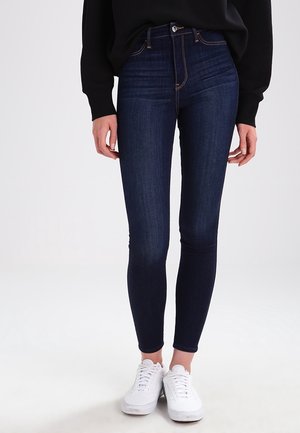 Woman wearing dark blue skinny jeans, black long-sleeve top, and white sneakers standing against a plain light background.