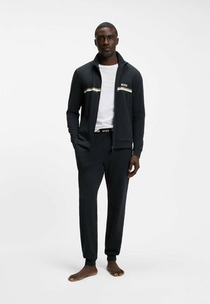 Uomo in piedi a piedi nudi, indossa una giacca nera con cerniera e strisce bianche, una maglietta bianca e pantaloni neri con il logo BOSS sulla giacca e sulla fascia della vita.