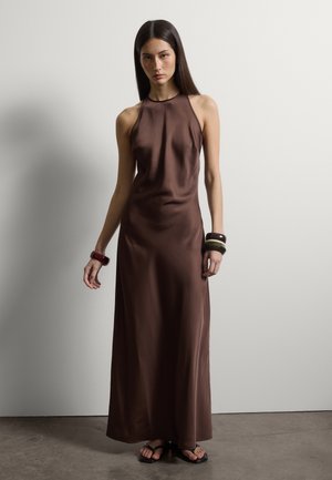 Mujer con vestido maxi de satén marrón sin mangas y cuello halter, con brazaletes negros y marrones y sandalias negras, de pie contra una pared lisa.