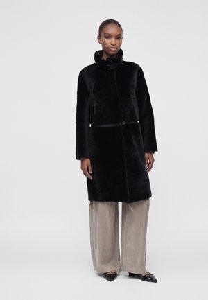 Femme portant un manteau en fausse fourrure noire avec un col montant et un pantalon beige à jambes larges, debout devant un fond uni.