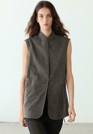 Femme aux cheveux longs et ondulés porte un gilet sans manches gris foncé boutonné sur un pantalon noir, debout devant un fond blanc uni.