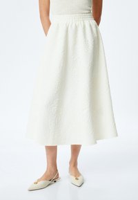 Jupe midi blanche avec un motif floral texturé, ceinture élastique et silhouette en A, associée à des chaussures à talons ouverts beige avec une accent doré.