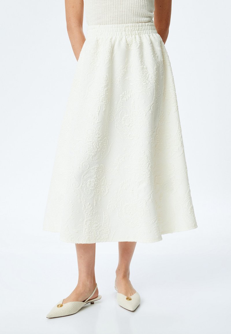 Jupe midi blanche avec un motif floral texturé, ceinture élastique et silhouette en A, associée à des chaussures à talons ouverts beige avec une accent doré.