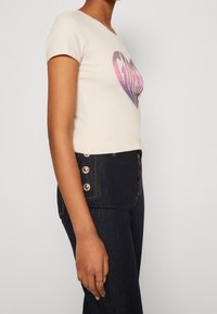 Camiseta corta de manga corta de color beige claro con un diseño de corazón en tonos rosa y púrpura. Llevada con unos jeans de mezclilla oscuros de tiro alto con detalles de botones dorados.