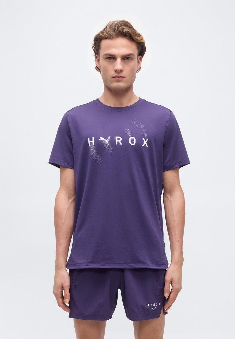 Νεαρός άνδρας που φοράει μωβ μπλουζάκι Puma Hyrox και αντίστοιχα σορτς, στέκεται μπροστά σε απλό φωτεινό φόντο.