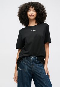 Görög szőke hajú fiatal nő fekete logós pólóban és laza sötétkék farmernadrágban, egyszínű világos háttér előtt áll.