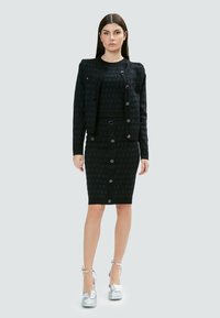 Abbigliamento coordinato nero composto da una gonna aderente e un giubbotto con bottoni, entrambi in un tessuto texturizzato con bottoni a contrasto e una maglietta abbinata.