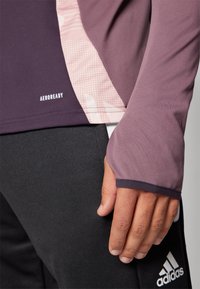 Lila langärmliges Sportoberteil mit pinken Musterelementen und dem Label "AEROREADY". Es hat eine enganliegende Passform und Daumenlöcher, kombiniert mit schwarzen Hosen.
